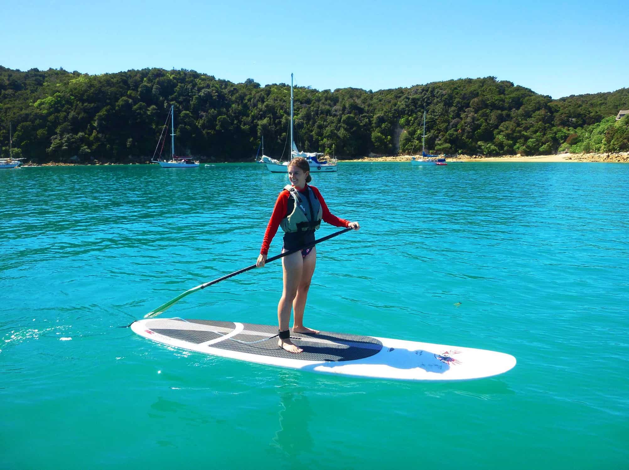 Woman standing on a paddleboard in a bay in New Zealand, with four small yachts anchored in the background.