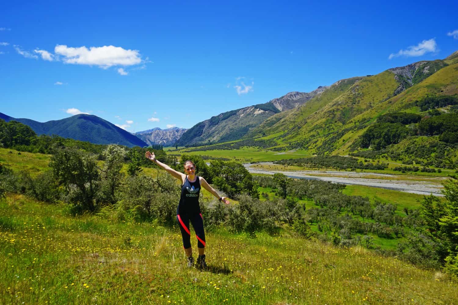 Author in hiking attire on a hillside in New Zealand, with a braided gravel river in the valley beyond and more large hills on the other side. Spring flowers are visible on the hills.