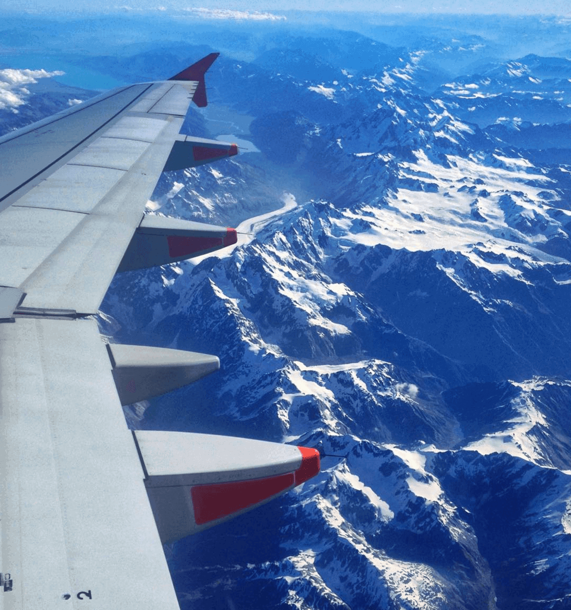 View of a plane wing as it flies over a mountain range in New Zealand, with snow-capped peaks and a river flowing through a valley.